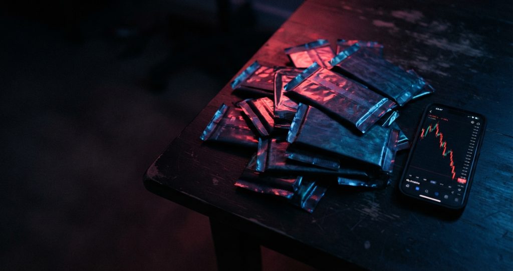 Pile de boosters de cartes scellés sur une table noire, éclairage néon rouge et bleu, à côté d'un téléphone affichant une courbe de prix qui s'effondre
